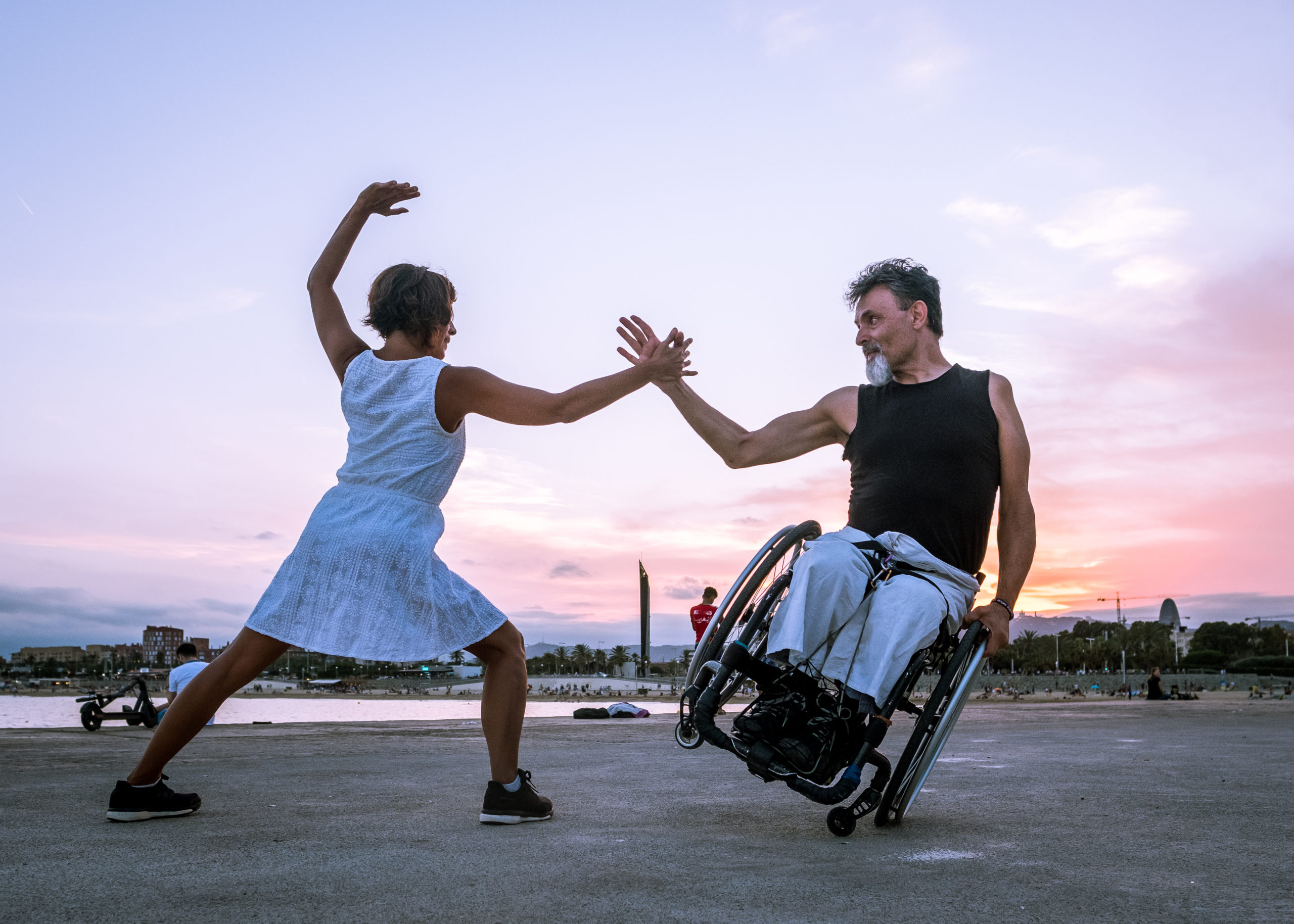 Donna con vestito bianco in piedi che tende la mano ad un uomo in sedia a rotelle in equilibrio su una sola ruota