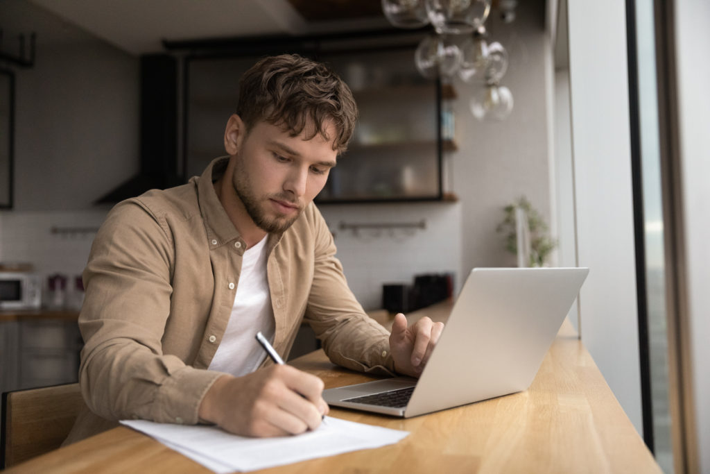 Ragazzo seduto a un tavolo che scrive su un foglio a fianco ad un computer portatile